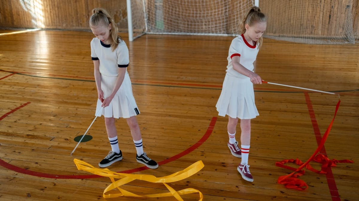 Two girls practice rhythmic gymnastics with ribbons on a wooden indoor court.